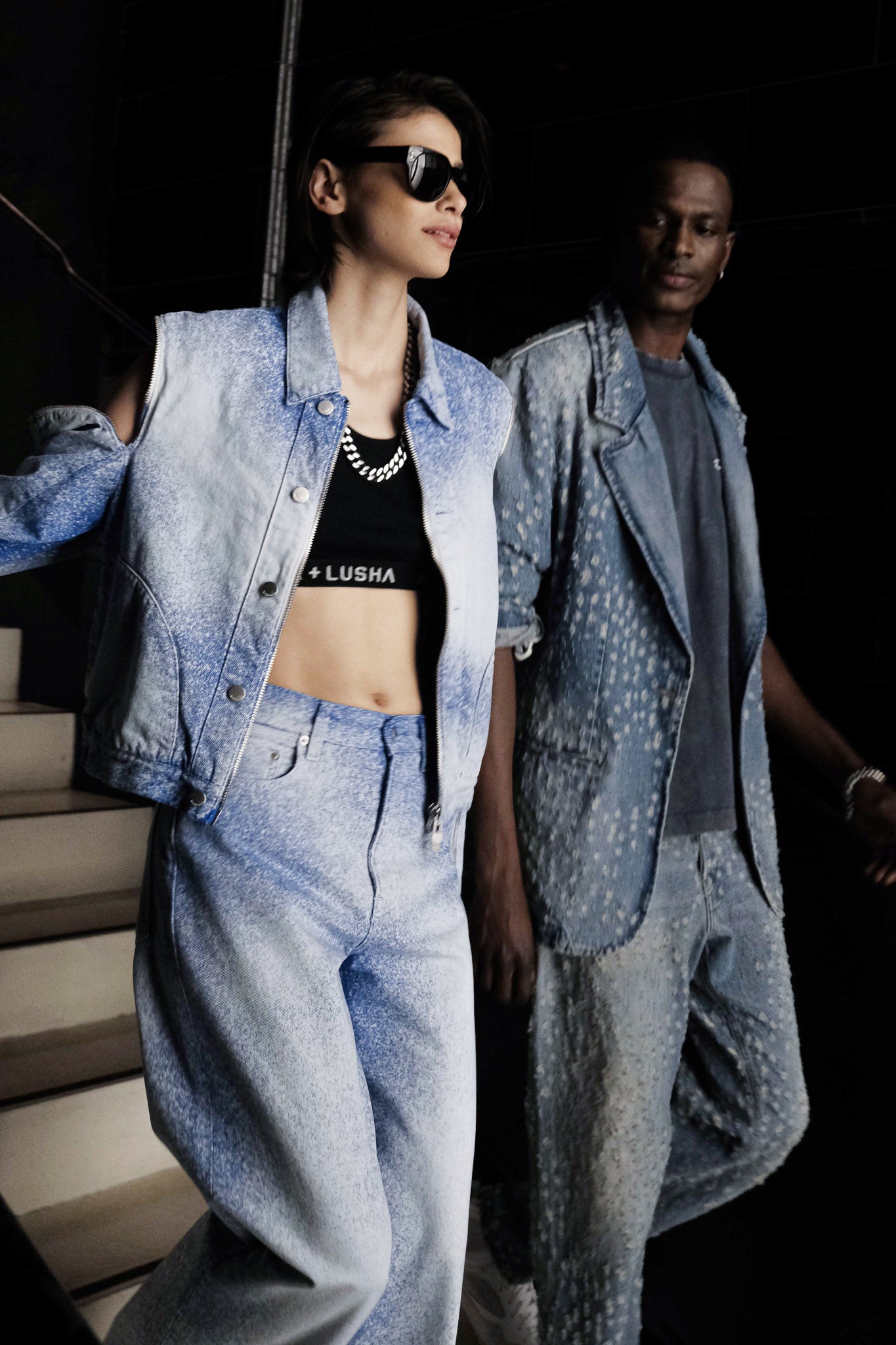 Female model wearing a blue ombre denim jacket and pants with a black K+LUSHA crop top, walking with a male model in a blue denim blazer with a unique spotted pattern and matching denim pants from K+, both models descending a staircase.